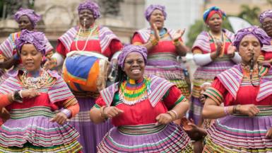 Women in traditional, multi-coloured Basotho outfits sing and dance 