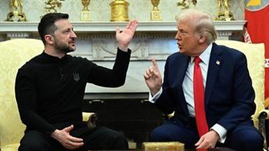 Zelensky in black, sitting on gold chair in front of gold trophies, holds up hand and faces Trump, who also sits on a gold chair and who has his forefinger raised while speaking