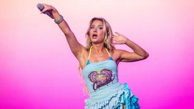 Zara Larsson performs on stage and she is holding a microphone outstretch to the crowd. She wears a blue sparkly dress.