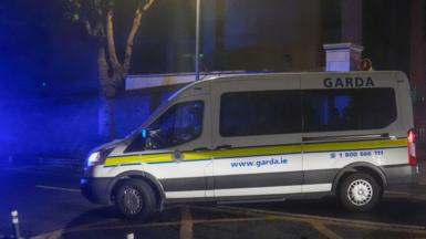A white Garda van with a yellow stripe along its side pulls out onto a main road on a dark night.