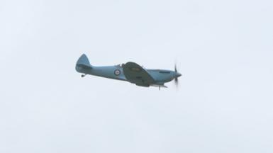 A Spitfire painted in a pale blue colour with a circular logo on the side flies through the air.