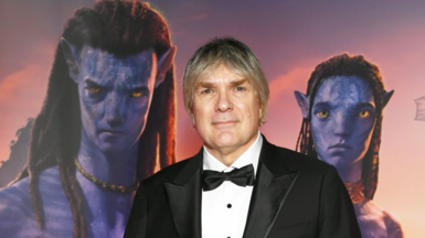 The image captures a formal moment at a promotional event for the movie Avatar. Composer Simon Franglen is dressed in a classic black tuxedo with a bow tie standing in front of a vibrant backdrop. The backdrop features two iconic Na'vi characters from the Avatar franchise—blue-skinned, yellow-eyed, and adorned with tribal markings—set against a warm, glowing background. The image was taken at a red carpet event / possibly a film premiere.