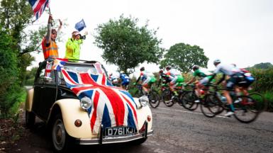 Fans wave flags in a car draped in a union jack as riders go past