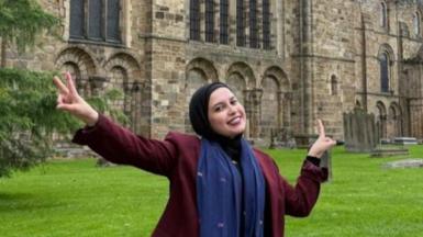 Sana el-Azab is sitting on a wall circling Durham Cathedral. She is smiling and is doing a peace sign with her hands.
