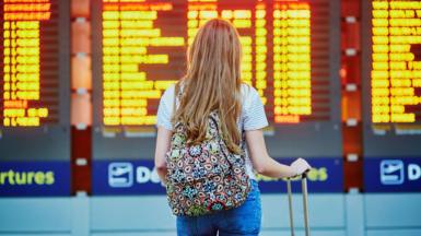A woman stares at a flight departures board. She has long blonde hair and has a rucksack on her back and a carry on case in her hand.