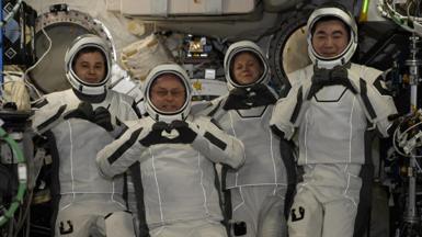 Four astronauts wearing white spacesuits and helmets pose for cameras smiling and making heart shapes with their hands and fingers. They are surrounded by wires and other equipment inside the International Space Station.