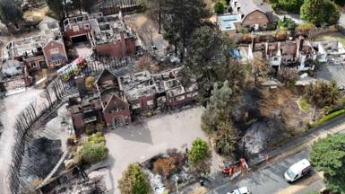 An aerial image shows several houses destroyed by fire, with their roofs gone and wreckage visible within. There is a line of dead trees next to the houses and areas of scorched earth. A fire engine is parked by one of the houses.