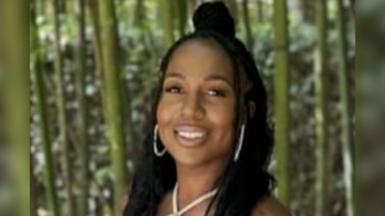 A young woman wearing gold jewellery smiles. She is standing in front of a forest.