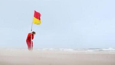 Lifeguard securing a red and yellow flag on a deserted beach with sand whipped up by strong winds 