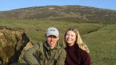 A man and a woman smiling sat in a barren field. He is wearing a grey hat with a green jacket. She's wearing a burgundy turtleneck.