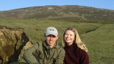 A man and a woman smiling sat in a barren field. He is wearing a grey hat with a green jacket. She's wearing a burgundy turtleneck.