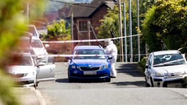 A forensic officer stood beside a blue car, the road is taped off with multiple white cars parked either side