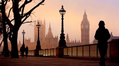 Westminster and Big Ben in the background from the other side of the river Thames at dusk. A moody figure in the foreground walks along the river