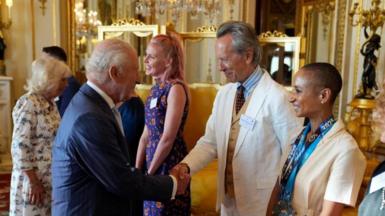 King Charles shakes hands with Richard E Grant at a reception at Buckingham Palace. Adele Roberts stands to Grant's left. Queen Camilla is greeting guests behind the King.