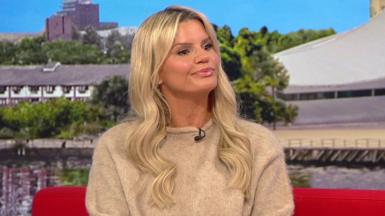 Kerry Katona sat on the BBC Breakfast red sofa. She has long blonde hair and wears a beige jumper