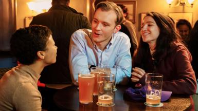 Three young people, two women, one man, are sat round a pub table with pints of beer. They are mid-conversation and smiling