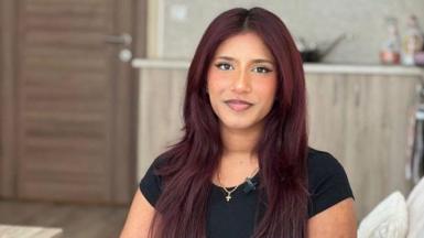 Freya wears a black t-shirt and sit on a cream couch. She has light burgundy-coloured hair. 