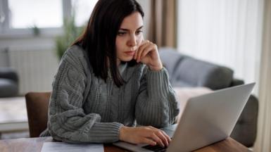 Woman with long dark hair and wearing a grey jumper sits at a desk looking at a laptop computer