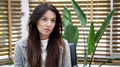 Zoe Trafford has long brown hair and wears a grey blazer and white top underneath. She is sitting against a background of wooden blinds against windows. The leaves of an indoor plant are also there.