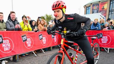 A man in black cycling gear, a red helmet and a red nose is leaning forward on a red tandem bike. There are red barriers in the street which read "Comic Relief Red Nose Day" with people stood behind them taking photographs on their phones.