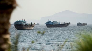 Boats atop the water pictured in a view of the Strait of Hormuz on a hazy day in mid-May with rock and plantation bokeh in the foreground and mountains in the background, taken from Oman on 22 May.