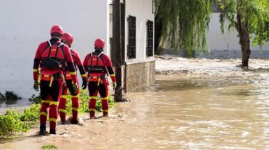 Three rescue workers wearing red and yellow wetsuits with red hard hats walk through ankle-deep brown water on a Spanish street.