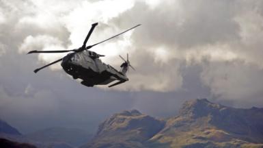 An RAF helicopter above a mountain range - it flies low to the cliff face against a dramatic grey cloud.