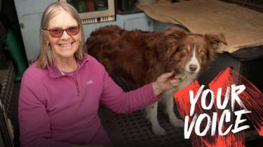 A woman in a pink fleece and wearing glasses sits in the back of a van with her border collie. 