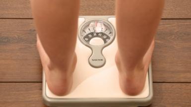 A generic image of a person standing on a pair of silver weight scales. You can see their bare legs and feet.