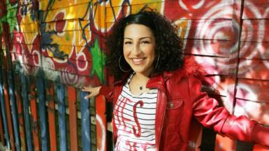A young woman with curly brown hair, wearing a red leather jacket and white-striped top, smiling at the camera and standing in front of a brightly graffitied wall