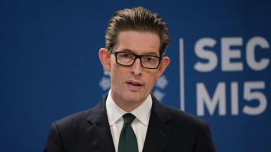 MI5 Director General Ken McCallum giving his speech. Behind him is a blue MI5 branded backdrop. He is wearing glasses, with a dark green tie and a suit, with his hair combed back.