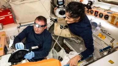 Butch is wearing a blue uniform and goggles. He appears to be fixing something and is wearing blue gloves. Suni is to positioned to Butch's right looking over his shoulder and watching. She has the same blue uniform but no goggles or gloves.