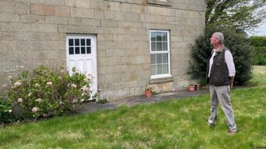 Martin Beal is looking at his house which is a Georgian stone built farmhouse with a white door and windows. He is a man, about 60 years of age, wearing a waist coat, white shirt and light coloured trousers.