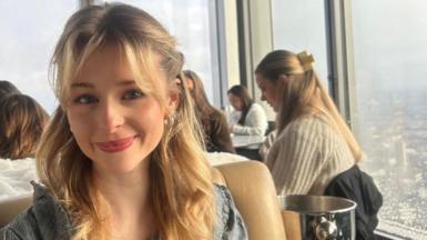 Eve Dulcie sits inside a in a rooftop restaurant with a skyline in the background. She smiles at the camera. She has blonde mid length hair, which is half pulled back from her face with her bangs loose in her face. 
