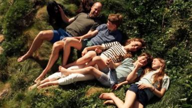 The Prince of Wales lies in the grass in a khaki tshirt and blue shorts, his hand is resting on his black spaniel dog, next to him is Prince George in a blue tshirt and green shorts, the Princess of Wales is smiling next to them in a white and navy stripped tshirt and white trousers, she holds the hand of Prince louis who wears a green shirt and chequered blue shorts, Princess charlottes is wearing black shorts and a pale green tshirt.