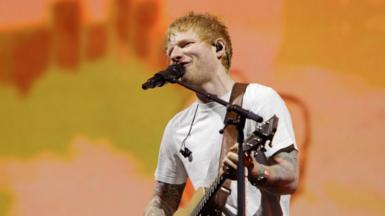 Ed Sheeran in a white shirt playing the guitar with a hazy orange background behind him