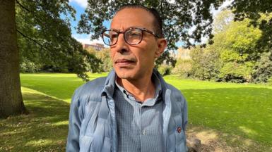 Dr Mohammed Abu Mughaisib, a medic evacuated from Gaza, stands in a park for a posed photograph. He is looking away to the left of the shot and has a blue/grey jacket and shirt on. He is wearing glasses. In the background are green bushes and trees.