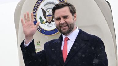 US Vice President JD Vance waves as he stands in front of the doors to an aircraft with "Vice President of the United States" written on it.