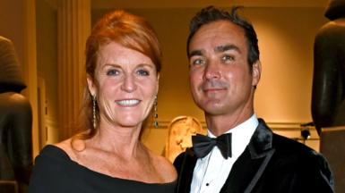 Sarah Ferguson in black dress with her hair up stands next to Manuel Fernandez who wears a black velvet jacket, white shirt and bow tie. They are both smiling at the camera. 