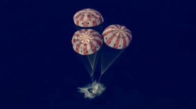 Three red and white parachutes bring the Orion capsule gently into the Pacific Ocean with a splash.