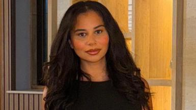 A woman wearing a black outfit stands indoors against a softly lit background with shelves and warm lighting.
