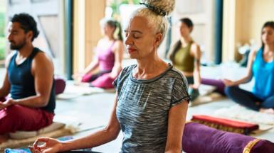 Stock photo shows a person sittting on the floor of a yoga studio while practising meditation with others in the background.