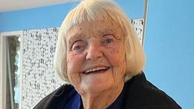 Jenny Nias, smiling, with white coloured bob hair, wearing a black coat.