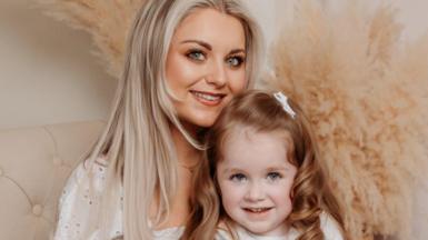 Hollie pictured on a cream chair with her mum Ann, the photo is professional. Hollie has long curly red/brown hair and is wearing a blue and white dress with a white cardigan. Ann has long blonde hair and is holding Hollie. 