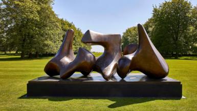 Large curved bronze sculpture by Henry Moore on a plinth, surrounded by open grass and trees in bright sunlight