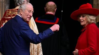 The King and Queen turn around to look behind them. The King holds a hand up while the Queen smiles