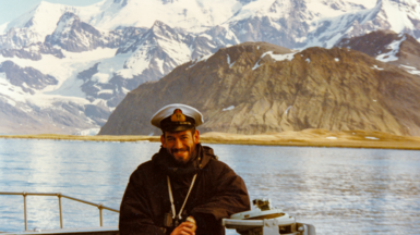 Commander Woods standing on a ship smiling with mountains behind him wearing a navy cap