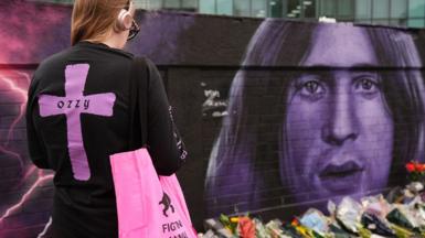A young woman with a large purple cross and the word Ozzy on the back of her black top, facing a purple mural of Ozzy's face, with a line of flowers below