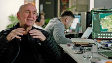Peter Molyenux is sitting in an office wearing a black jacket and black top - behind him a man with white headphones in his staring and his computer screen. Next to Peter is part of a computer screen showing Masters of Albion. The desk has notebooks and a glass of water.
