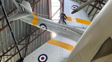 A person attached to a rope system, stands on an RAF plane that's hanging from the ceiling of a hangar.